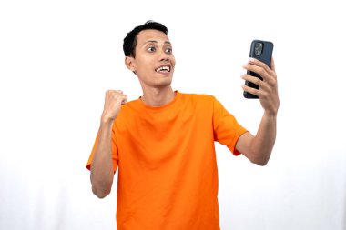 Turuncu bir tişört giyen Asyalı genç bir adamın portresi mutlu bir ifadeyle akıllı telefondan mutlu haberler almasına şaşırdı. Beyaz arkaplanda izole.