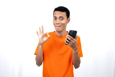 Turuncu tişört giyen genç bir Asyalı 'nın portresi tamam işareti veriyor, kabul ediyorum, elinde akıllı telefon tutarken. Beyaz arkaplanda izole.
