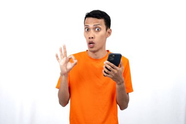 Turuncu tişört giyen genç bir Asyalı 'nın portresi tamam işareti veriyor, kabul ediyorum, elinde akıllı telefon tutarken. Beyaz arkaplanda izole.