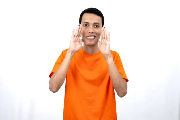 stock image Portrait of a young Asian man wearing an orange t-shirt with his hand near his mouth whispering gossip and telling secrets while sharing news isolated on white background.