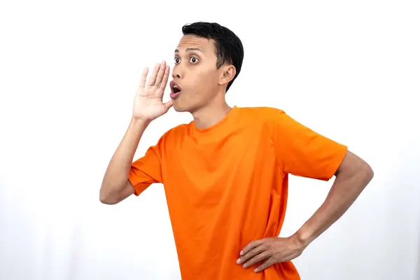 stock image Portrait of a young Asian man wearing an orange t-shirt with his hand near his mouth whispering gossip and telling secrets while sharing news isolated on white background.