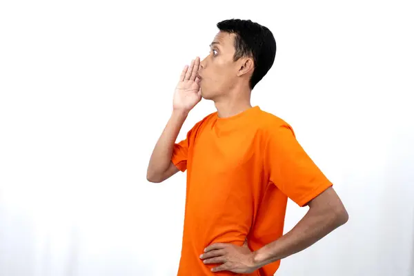 stock image Portrait of a young Asian man wearing an orange t-shirt with his hand near his mouth whispering gossip and telling secrets while sharing news isolated on white background.