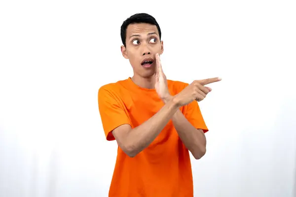 stock image Portrait of a young Asian man wearing an orange t-shirt with his hand near his mouth in a whispering pose while pointing to the side of the copy space. Isolated on white background.