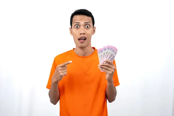 Yüzbinlerce rupiah taşırken mutlu, şaşırmış ve mutlu bir ifadeye sahip turuncu bir tişört giyen Asyalı genç bir adamın portresi. beyaz arkaplanda izole edilmiş.