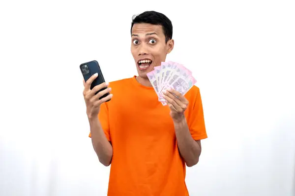 Bir akıllı telefon ve yüz binlerce rupiah taşırken mutlu, şaşkın ve memnun bir ifadeye sahip turuncu bir tişört giyen genç bir Asyalı 'nın portresi. beyaz arkaplanda izole edilmiş.