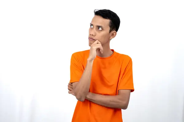 stock image Portrait of a young Asian man wearing an orange t-shirt posing thinking, searching for ideas, or confused. Isolated on white background.