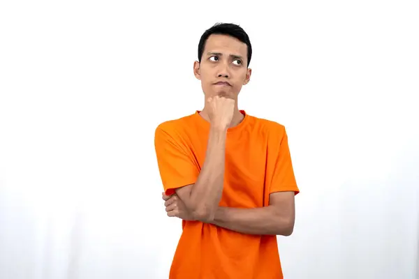 stock image Portrait of a young Asian man wearing an orange t-shirt posing thinking, searching for ideas, or confused. Isolated on white background.