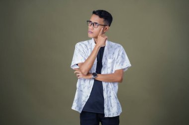 Gözlüklü, beyaz gömlekli, düşünceli, başı dönen ya da fikir arayan Asyalı bir adam. Ordu yeşil arka planında izole edilmiş..