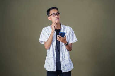 Gözlüklü genç Asyalı adam kafası karışık bir ifade sergiliyor, akıllı telefon tutarken düşünüyor. Ordu yeşil arka planında izole edilmiş..