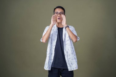 Heyecanlı, şok olmuş ya da heyecanlı bir poz. Ağızdan ağıza bağırıyordu. Gözlüklü, sıradan beyaz tişörtlü Asyalı bir adam askeri yeşil arka planda izole edilmiş...