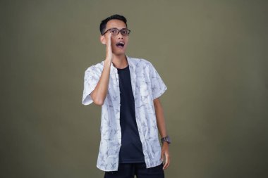 Heyecanlı, şok olmuş ya da heyecanlı bir poz. Ağızdan ağıza bağırıyordu. Gözlüklü, sıradan beyaz tişörtlü Asyalı bir adam askeri yeşil arka planda izole edilmiş...