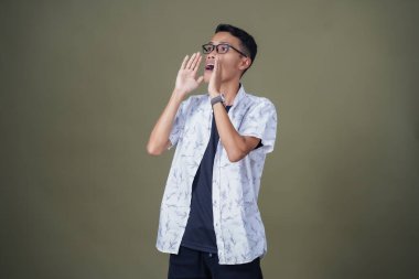 Heyecanlı, şok olmuş ya da heyecanlı bir poz. Ağızdan ağıza bağırıyordu. Gözlüklü, sıradan beyaz tişörtlü Asyalı bir adam askeri yeşil arka planda izole edilmiş...