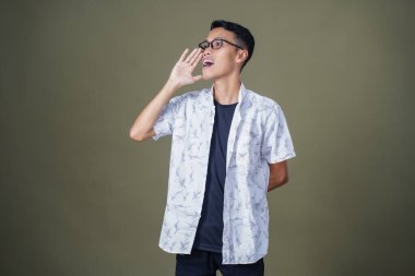 Heyecanlı, şok olmuş ya da heyecanlı bir poz. Ağızdan ağıza bağırıyordu. Gözlüklü, sıradan beyaz tişörtlü Asyalı bir adam askeri yeşil arka planda izole edilmiş...