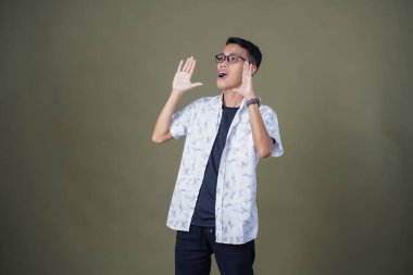 Heyecanlı, şok olmuş ya da heyecanlı bir poz. Ağızdan ağıza bağırıyordu. Gözlüklü, sıradan beyaz tişörtlü Asyalı bir adam askeri yeşil arka planda izole edilmiş...