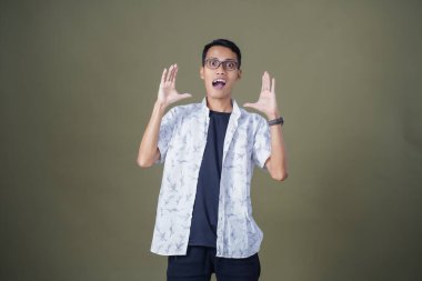 Heyecanlı, şok olmuş ya da heyecanlı bir poz. Ağızdan ağıza bağırıyordu. Gözlüklü, sıradan beyaz tişörtlü Asyalı bir adam askeri yeşil arka planda izole edilmiş...