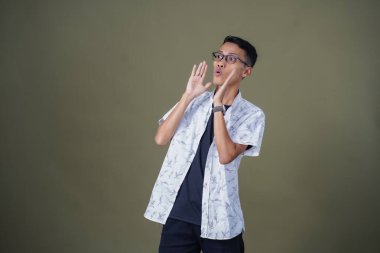 Heyecanlı, şok olmuş ya da heyecanlı bir poz. Ağızdan ağıza bağırıyordu. Gözlüklü, sıradan beyaz tişörtlü Asyalı bir adam askeri yeşil arka planda izole edilmiş...