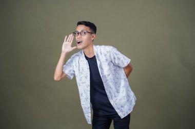 Heyecanlı, şok olmuş ya da heyecanlı bir poz. Ağızdan ağıza bağırıyordu. Gözlüklü, sıradan beyaz tişörtlü Asyalı bir adam askeri yeşil arka planda izole edilmiş...