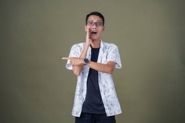 Heyecanlı, şok olmuş ya da heyecanlı bir poz. Boş uzayın kenarını gösteren el işaretleriyle bağırıyorlar. Gözlüklü, sıradan beyaz tişörtlü Asyalı bir adam askeri yeşil arka planda izole edilmiş..