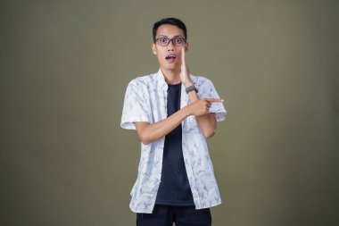 Heyecanlı, şok olmuş ya da heyecanlı bir poz. Boş uzayın kenarını gösteren el işaretleriyle bağırıyorlar. Gözlüklü, sıradan beyaz tişörtlü Asyalı bir adam askeri yeşil arka planda izole edilmiş..