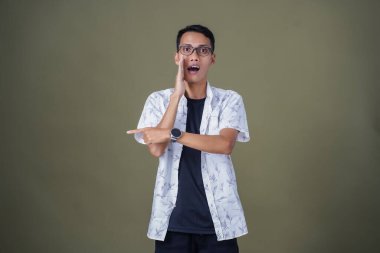 Heyecanlı, şok olmuş ya da heyecanlı bir poz. Boş uzayın kenarını gösteren el işaretleriyle bağırıyorlar. Gözlüklü, sıradan beyaz tişörtlü Asyalı bir adam askeri yeşil arka planda izole edilmiş..