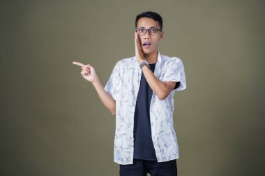 Heyecanlı, şok olmuş ya da heyecanlı bir poz. Boş uzayın kenarını gösteren el işaretleriyle bağırıyorlar. Gözlüklü, sıradan beyaz tişörtlü Asyalı bir adam askeri yeşil arka planda izole edilmiş..