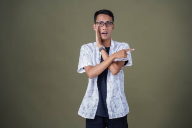 Heyecanlı, şok olmuş ya da heyecanlı bir poz. Boş uzayın kenarını gösteren el işaretleriyle bağırıyorlar. Gözlüklü, sıradan beyaz tişörtlü Asyalı bir adam askeri yeşil arka planda izole edilmiş..