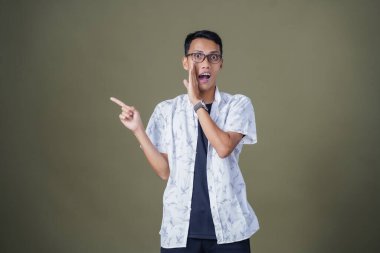 Heyecanlı, şok olmuş ya da heyecanlı bir poz. Boş uzayın kenarını gösteren el işaretleriyle bağırıyorlar. Gözlüklü, sıradan beyaz tişörtlü Asyalı bir adam askeri yeşil arka planda izole edilmiş..