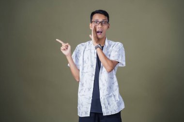 Heyecanlı, şok olmuş ya da heyecanlı bir poz. Boş uzayın kenarını gösteren el işaretleriyle bağırıyorlar. Gözlüklü, sıradan beyaz tişörtlü Asyalı bir adam askeri yeşil arka planda izole edilmiş..