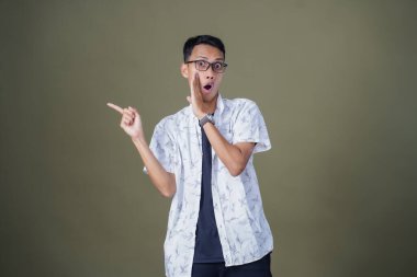 Heyecanlı, şok olmuş ya da heyecanlı bir poz. Boş uzayın kenarını gösteren el işaretleriyle bağırıyorlar. Gözlüklü, sıradan beyaz tişörtlü Asyalı bir adam askeri yeşil arka planda izole edilmiş..