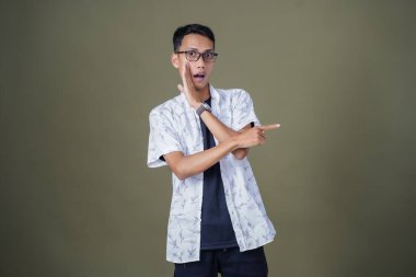 Heyecanlı, şok olmuş ya da heyecanlı bir poz. Boş uzayın kenarını gösteren el işaretleriyle bağırıyorlar. Gözlüklü, sıradan beyaz tişörtlü Asyalı bir adam askeri yeşil arka planda izole edilmiş..