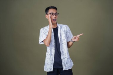 Heyecanlı, şok olmuş ya da heyecanlı bir poz. Boş uzayın kenarını gösteren el işaretleriyle bağırıyorlar. Gözlüklü, sıradan beyaz tişörtlü Asyalı bir adam askeri yeşil arka planda izole edilmiş..