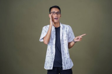 Heyecanlı, şok olmuş ya da heyecanlı bir poz. Boş uzayın kenarını gösteren el işaretleriyle bağırıyorlar. Gözlüklü, sıradan beyaz tişörtlü Asyalı bir adam askeri yeşil arka planda izole edilmiş..