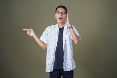 Heyecanlı, şok olmuş ya da heyecanlı bir poz. Boş uzayın kenarını gösteren el işaretleriyle bağırıyorlar. Gözlüklü, sıradan beyaz tişörtlü Asyalı bir adam askeri yeşil arka planda izole edilmiş..
