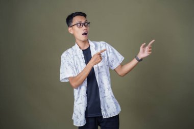 Heyecanlı, şok olmuş ya da heyecanlı bir poz. Boş uzayın kenarını gösteren el işaretleriyle bağırıyorlar. Gözlüklü, sıradan beyaz tişörtlü Asyalı bir adam askeri yeşil arka planda izole edilmiş..