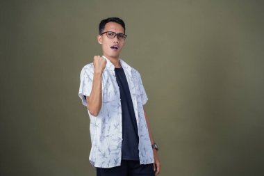 Sıradan gözlüklü genç Asyalı adam yumruğunu sıkarak poz veriyor, zafer ya da güçlü bir tarz. Ordu yeşil arka planında izole edilmiş..