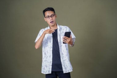 Gündelik gözlüklü genç Asyalı adam akıllı telefonu işaret ederken şaşırdı. Ordu yeşil arka planında izole edilmiş..