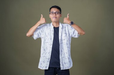 Gözlüklü, beyaz gömlekli, Asyalı bir adam. Mutlu bir şekilde baş parmak hareketi yapmak. Ordu yeşil arka planında izole edilmiş..