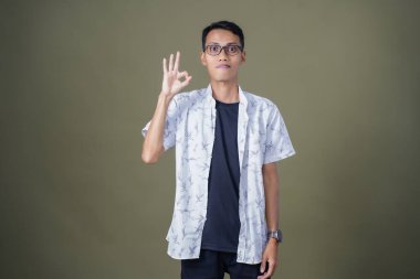 Gözlüklü genç Asyalı adam akıllı telefonu tutarken iyi işareti gösteriyor. Ordu yeşil arka planında izole edilmiş..