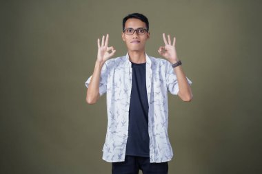 Gözlüklü genç Asyalı adam akıllı telefonu tutarken iyi işareti gösteriyor. Ordu yeşil arka planında izole edilmiş..