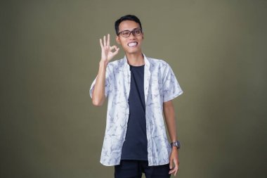 Gözlüklü genç Asyalı adam akıllı telefonu tutarken iyi işareti gösteriyor. Ordu yeşil arka planında izole edilmiş..