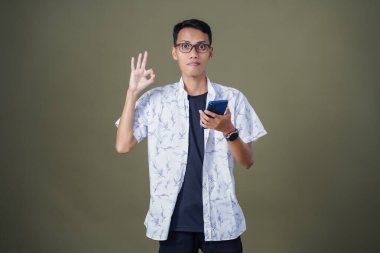 Gözlüklü genç Asyalı adam akıllı telefonu tutarken iyi işareti gösteriyor. Ordu yeşil arka planında izole edilmiş..