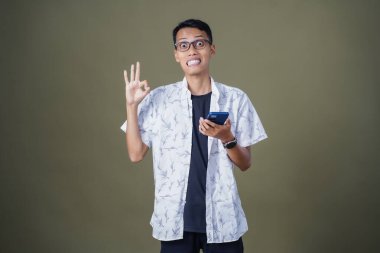 Gözlüklü genç Asyalı adam akıllı telefonu tutarken iyi işareti gösteriyor. Ordu yeşil arka planında izole edilmiş..