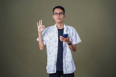 Gözlüklü genç Asyalı adam akıllı telefonu tutarken iyi işareti gösteriyor. Ordu yeşil arka planında izole edilmiş..