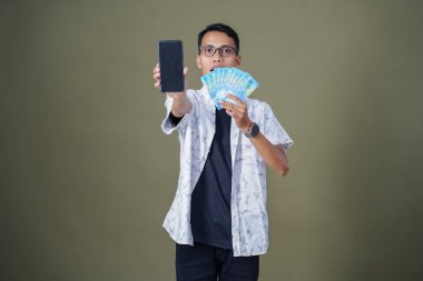 Gözlüklü genç Asyalı adam mutlu ve şaşırmış bir ifade sergiliyor, bir sürü fatura taşıyor ve elinde akıllı telefon tutuyor. Ordu yeşil arka planında izole edilmiş..