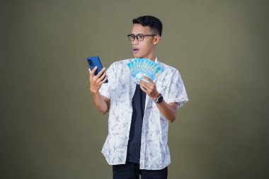 Gözlüklü genç Asyalı adam mutlu ve şaşırmış bir ifade sergiliyor, bir sürü fatura taşıyor ve elinde akıllı telefon tutuyor. Ordu yeşil arka planında izole edilmiş..