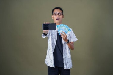 Gözlüklü genç Asyalı adam mutlu ve şaşırmış bir ifade sergiliyor, bir sürü fatura taşıyor ve elinde akıllı telefon tutuyor. Ordu yeşil arka planında izole edilmiş..