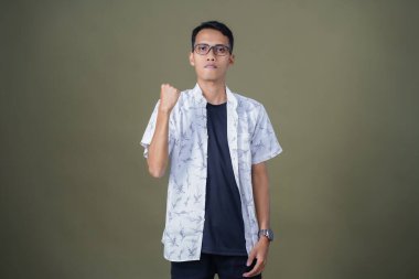 Sıradan gözlüklü genç Asyalı adam yumruğunu sıkarak poz veriyor, zafer ya da güçlü bir tarz. Ordu yeşil arka planında izole edilmiş..