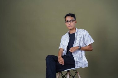 Gözlüklü genç Asyalı adam rahat rahat oturuyor. Ordu yeşil arka planında izole edilmiş..