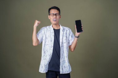 Gündelik gözlüklü genç Asyalı adam akıllı telefonu işaret ederken şaşırdı. Ordu yeşil arka planında izole edilmiş..