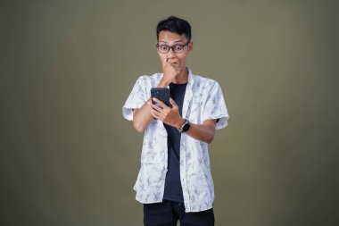 Gündelik gözlüklü genç Asyalı adam akıllı telefonu işaret ederken şaşırdı. Ordu yeşil arka planında izole edilmiş..